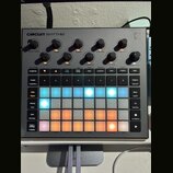 Šedý sampler Novation Circuit Rhythm s mřížkou podsvícených padů v modrých a oranžových barvách. V horní části jsou umístěny ovládací knoflíky s popi…
