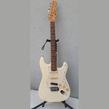 Bílá elektrická kytara typu Stratocaster s hnědým hmatníkem a krčkem z přírodního dřeva. Na hlavě je svisle umístěné logo "LAVA MUSIC". Kytara má tři…