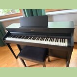 Černý digitální piano v podobě konzole s přiloženým polstrovaným černým stolečkem. Na přední horní hraně je umístěn nápis Roland. Nástroj má standard…