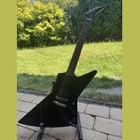 Černá elektrická kytara s ostře hranatým tělem typu Explorer umístěná na černém stojanu. Na hlavě hmatníku je bílý nápis Gibson. Kytara má dva humbuc…