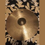 Zlatavý kovový činel s viditelným kovánívým povrchem a klepaným středem. V horní části je černý nápis HH, pod ním text Rare Bell Dry Ride a dále 21"/…
