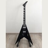 Černá elektrická kytara s tvarem V umístěná na stojanu. Na hlavě hmatníku je bílý nápis Jackson. Kytara má dva humbuckery, ladicí kolíky na hlavě a b…