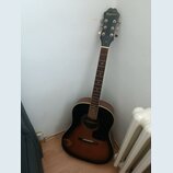 Akustická kytara s tmavým korpusem v provedení sunburst. Na hlavě hmatníku je bílý nápis Epiphone. Nástroj je opřen o bílou zeď vedle radiátoru.