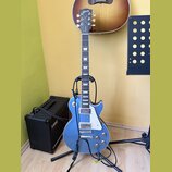 Modrá elektrická kytara typu Les Paul s dvěma humbuckery a bílým pickguardem, umístěná na černém stojanu. Na hlavě hmatníku je nápis Gibson a modelov…