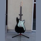 Černá elektrická kytara s bílým pickguardem umístěná na černém stojanu. Na hlavě hmatníku je viditelné logo Fender. Nástroj má tři snímače typu singl…