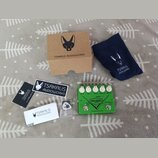 Sada příslušenství a kytarový efekt v zelené barvě s nápisy RIFF RAFF MKII, BOOST a Tsakalis AudioWorks. Soubor obsahuje hnědou kartonovou krabici, t…