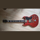 Červená elektrická kytara typu SG s tmavým hmatníkem a bílými vložkami. Na těle jsou dva humbuckery, čtyři oválné knofíky a přepínač. Na pravé straně…