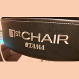 Detail černé polstrované sedací plochy z materiálu imitující kůži s bílým prošíváním. Na povrchu je vyšívka v bílé barvě s nápisy 1st CHAIR a TAMA. P…