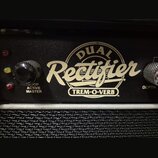 Detailní pohled na ovládací panel černého kytarového komba. Uprostřed je zlatý nápis DUAL Rectifier a pod ním v obdélníku TREM-O-VERB. Vlevo jsou tři…