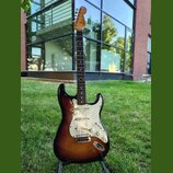 Elektrická kytara s tmelem v barvě sunburst, bílým pickguardem a hnedým hmatníkem. Na hlavě je viditelné logo Fender. Nástroj má tři snímače, pětipos…