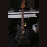 Černá akustická kytara s vyztřižnutým otvorem v korpusu a bílým lemováním. Na hlavě hmatníku je viditelné logo Fender. Nástroj stojí na stojanu před…