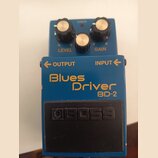 Modrý efekt pedál s nápisy Blues Driver a BD-2. Na spodním černém spínači je vyražené logo BOSS. Horní část obsahuje tři ovládací knoflíky s popisky…