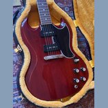 Elektrokytara s tmavě červeným lakovaným povrchem a černým pickguardem v hnědém plišovém kufru. Nástroj má dva humbuckery, stříbrný most a čtyři ovlá…