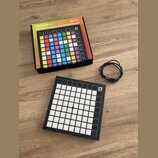 Na fotografii je černý MIDI kontrolér Novation Launchpad X s mřížkou bílých podsvícených polí a ovládacími tlačítky po stranách. Vedle zařízení leží…