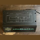 Černý elektronický modul s nápisy YAMAHA, FM EXPANDER a TX7 TONE GENERATOR. Horní kryt obsahuje schéma s texty DX SERIES, DATA ENTRY SLIDER, PROGRAM…