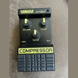 Černý kompresor s nápisem YAMAHA CO-10M.II v horní části a žlutým nápisem COMPRESSOR na spodním panelu. Zařízení má tři černé oválné knoflíky s žlutý…