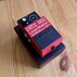 Červený kytarový pedál Loop Station RC-1 značky BOSS. Na horní části jsou nápisy INPUT A (MONO), INPUT B, OUTPUT A (MONO), OUTPUT B a LOOP. V dolní č…