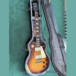 Elektrická kytara typu Les Paul s barevným přechodem od žluté k tmavě hnědé v černém tvrdém kufru s šedým polstrováním. Na hlavě hmatníku je bílý náp…