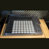 Černý MIDI kontrolér s mřížkou 8x8 bílých podsvícených padů. Zařízení je umístěno v kartonové krabici na dřevěném povrchu, v pozadí je vidět akustick…