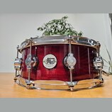 Červený snare drum s kovovým obsazem a lesklým povrchem. Uprostřed korpusu je bílý oválný štítek s nápisy drums, dw a DESIGN SERIES. Nástroj vykazuje…