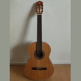 Klasická kytara s přírodním dřevěným povrchem a hnědým hmatníkem. Na hlavě nástroje je umístěno bílé písmeno A. Kytara má kulatý otvor s dekorativní…