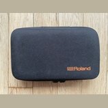 Černý obdélníkový pouzdro z textilie s texturovaným povrchem. V pravém dolním rohu je oranžové logo a nápis Roland. Pouzdro leží na dřevěném podkladu.