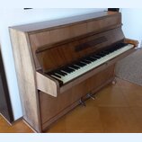 Hnědé dřevěné piano s klaviaturou. Nástroj vykazuje známky opotřebení, zejména v podobě odřenin a vybledlých míst na levém bočním panelu. Na klaviatu…