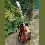 Polodupatá elektrická kytara s tmavě hnědým lakem v přechodu (sunburst) a zlatými prvky. Na hlavě je bílý nápis Epiphone. Nástroj má dva humbuckery,…