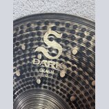 Detailní pohled na mosazný činel s tmavým, nepravidelným povrchem a viditelnými rýhami. Uprostřed je zlaté logo ve tvaru stylizovaného písmene S a po…