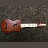 Hnědý dřevěný ukulele s hnedým korpusem a světlým hmatníkem. Uprostřed ozvučovacího otvoru je vidět štítek s nápisem "Kala". Nástroj vykazuje známky…