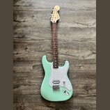 Světle zelená elektrická kytara typu Stratocaster s bílým perleťovým pickguardem a hnedým hmatníkem. Na hlavě je černý nápis Fender Stratocaster. Nás…