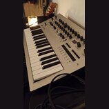 Stříbrný klaviatura synthesizer s bílými a černými klávesami. Na horní části jsou umístěny knoflíky, přepínače a tlačítka s nápisy jako PROGRAM VALUE…