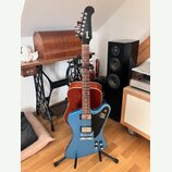 Modrá elektrická kytara s černým hmatníkem a hlavou, umístěná na černém stojanu. Na hlavě je bílý nápis Gibson. Tělo kytary má modrý lak, dva snímače…