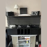 Na fotografii je set DJ vybavení umístěné na černých akustických panelech a bílém stolku. V horní části jsou dva černé studiové monitory, vprostřed č…