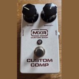Stříbrný efekt pedál s nápisy MXR CUSTOM SHOP a CUSTOM COMP. Na horní straně jsou dva černé oválné ovládací knoflíky s popisky OUTPUT a SENSITIVITY.…