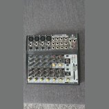 Šedý mixovací pult s nápisem XENYX 1202 a logem BEHRINGER. Zařízení obsahuje čtyři mikrofonní vstupy XLR, několik lineárních vstupů RCA a jack, poten…