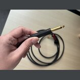 Ruka drží konec černého audio kabelu s pozlaceným 6,3mm jack konektorem. Na černém plastovém pouzdře konektoru je vyraženo logo NEUTRIK. V pozadí je…