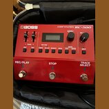 Červený looper Boss Loop Station RC-500 v černé brašně. Zařízení má digitální displej, několik ovládacích tlačítek a dva posuvníky. V dolní části jso…