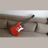 Elektrická kytara s červeným lakovaným korpusem a hnědým hmatníkem. Nástroj má dva humbuckery, bílý plech sylovací kryt a čtyři zlaté ovládací knoflí…