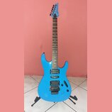 Modrá elektrická kytara s hrazdicem a dvěma humbuckery v pozici kobylníku a krku. Na hlavě hmatníku je bílý nápis Ibanez. Nástroj stojí na černém sto…