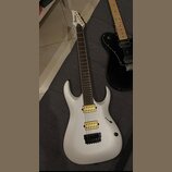 Bílá elektrická kytara s matným povrchem a hnědým hmatníkem. Na hlavě je nápis Ibanez, na těle jsou dva žluté humbuckery a černý kobylník. Vedle ní l…