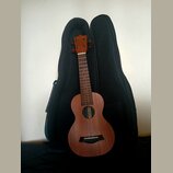 Hnědý dřevěný ukulele s černým pouzdrem v pozadí. Na hlavě nástroje i uvnitř ozvukové otvory je vidět nápis „conga“. Nástroj vykazuje známky běžného…