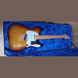 Elektrická kytara s t bodies v barevném přechodu od žluté po tmavě hnědou, umístěná v modrém polstrovaném kufru. Nástroj má bílý pickguard, zlaté har…