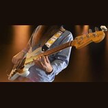 Bílá elektrická basová kytara s hnědým hmatníkem a světle dřevěnou hlavou. Na hlavě je vidět nápis Fender a text Jazz Bass. Nástroj má zlatý plech s…