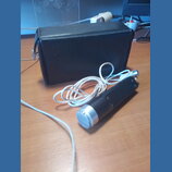 Na fotografii je černý mikrofon s kovovou stříbrnou mřížkou a bílým kabelem. Vedle mikrofonu je umístěno černé obdélníkové pouzdro z materiálu připom…