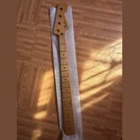 Dřevěný hmatník a hlava basové kytary v přirozené barvě dřeva, položené na bílém polystyrenovém podkladu. Na hlavě je černý nápis "Fender" a menší te…