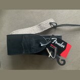 Černý kytarový popruh z nubukové kůže s logem Fender vyraženým do materiálu. K popruhu je připevněn červenobílý papírový štítek s nápisy Fender a Str…