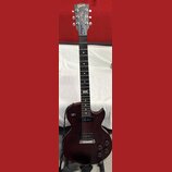 Tmavě červená elektrická kytara typu Les Paul s černým hmatníkem a dvěma humbuckery. Na hlavě je bílý nápis Gibson a pod ním kurzívou Les Paul Model.…