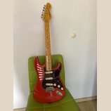 Elektrická kytara typu Stratocaster s červeným bodies, černým pickguardem a světle hnědým hnedem hmatníkem. Na hlavě je nápis "Vintage Modified Strat…