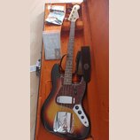 Elektrická basová kytara s trupem v barevnosti sunburst a hnědým hmatníkem, umístěná v otevřeném kufru s oranžovou výstuhou. Na hlavě je nápis Fender…